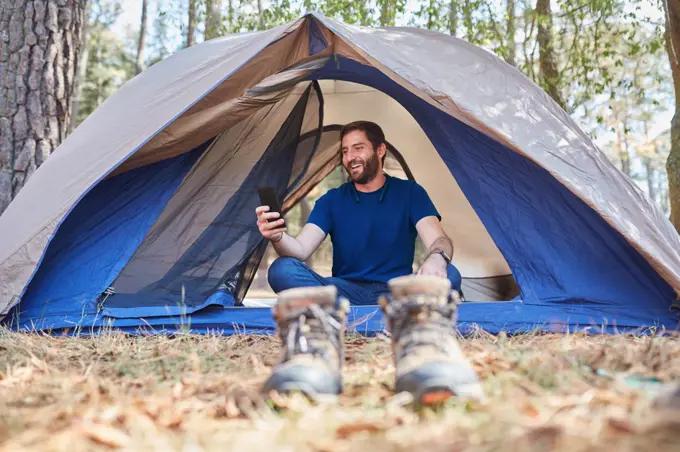 smiling young hispanic man using his cellphone sitting inside his tent