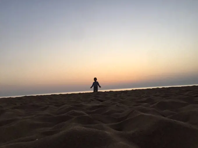 A little kid is standing on the beach with in sunset spreading hands