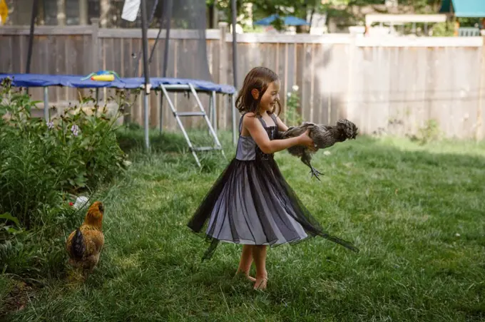 A little laughing girl holds a chicken and twirls in backyard garden