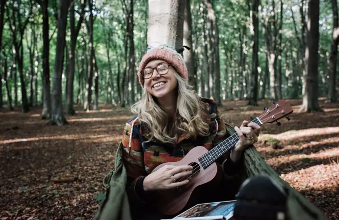 woman happily playing the ukulele in the forest on a hammock