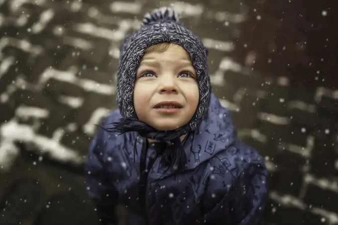 Small boy in blue winter clothes looking up at the snowing sky