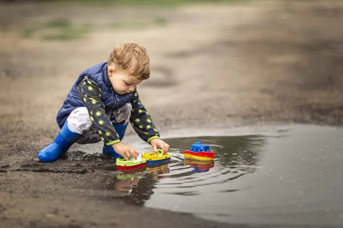 Boy in rain boots crouching in puddle and playing with ships