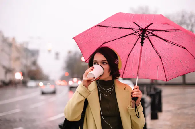 Young woman with an umbrella drinking coffee walking in city