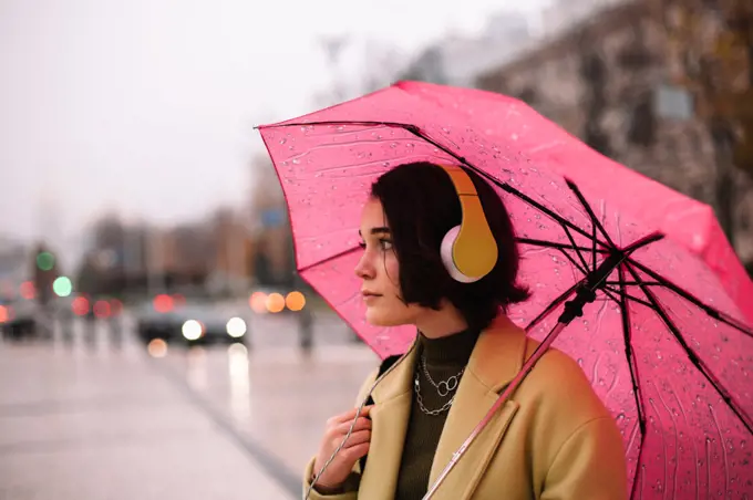 Thoughtful young woman in headphones holding umbrella standing in city