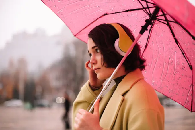 Thoughtful young woman in headphones holding umbrella walking in city