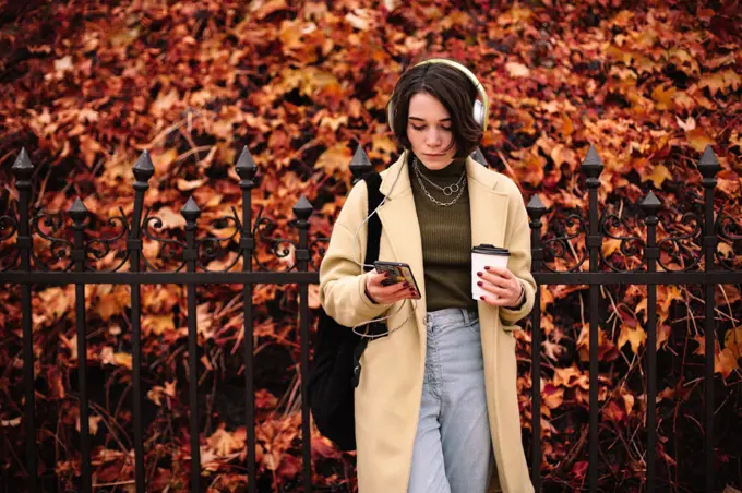 Young woman listening music in headphones standing in city in autumn