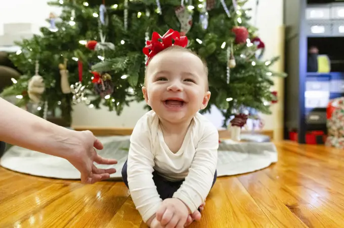 Happy Baby Sits on Floor Next to Christmas Tree with Bow on Head