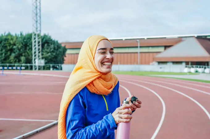 Joyful ethnic woman drinking water from bottle on racetrack