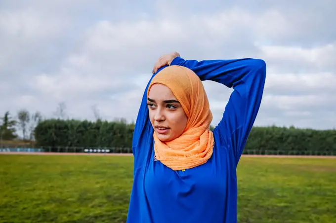 Muslim woman doing overhead tricep stretch on sports field