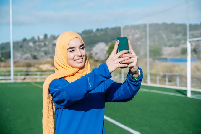 Happy ethnic woman taking selfie on smartphone on sports field