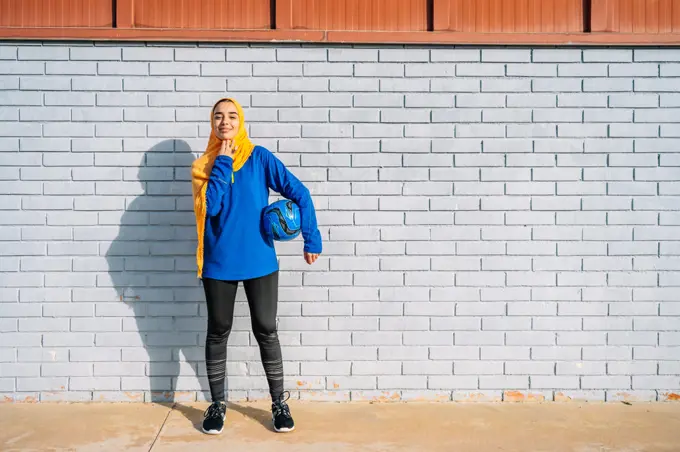 Smiling Arab female with football ball near brick building