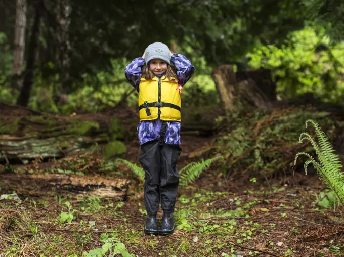 A young girl in rain gear and life jacket stands in the forest