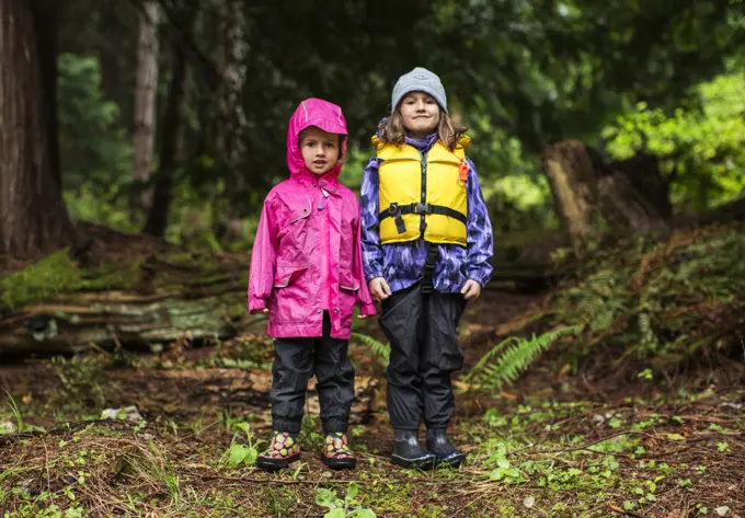 Two young girls in rain gear stand in a forest