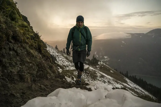 A man hikes up a mountain trail with snow and clouds