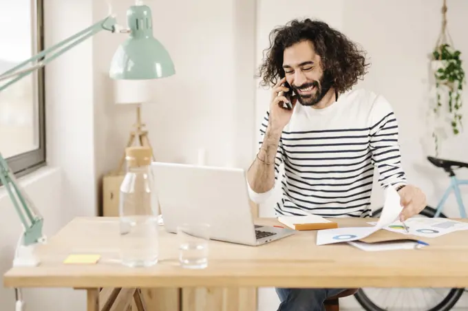 Cheerful young man talking on phone while working from home