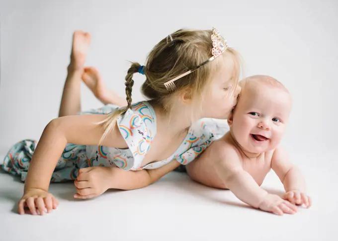 Sister kisses baby brother laying on white backdrop