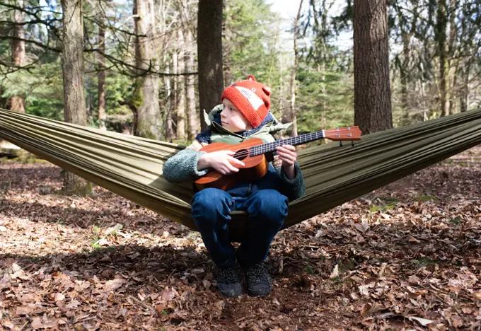 boy sat in a hammock in the forest playing the ukelele relaxing
