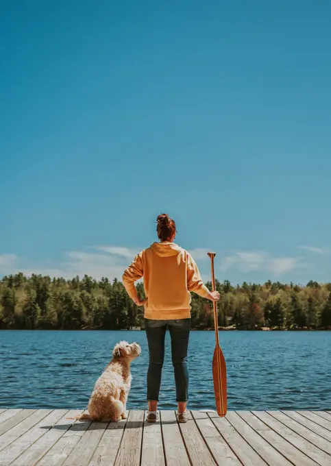Woman standing on dock with dog holding paddle looking out at lake.