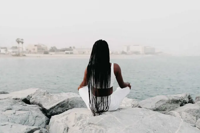 Woman with braids sittingon the rocks at the beach meditating
