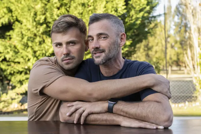 A young man hugging his husband outdoors.  Both are looking at camera