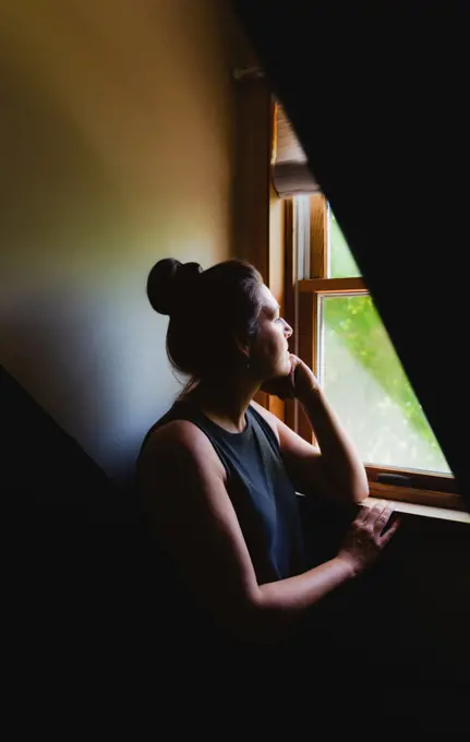 Woman looking out of a window in a dark room in the daytime.