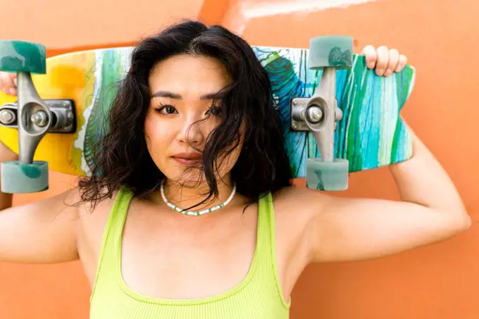 Asian woman standing next to the orange wall holding a skateboar