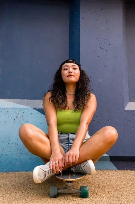 Asian woman sitting on her skateboard next to the blue wall 