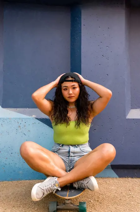 Asian woman sitting on her skateboard next to the blue wall 