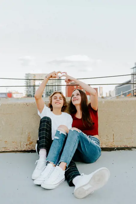 boyfriend and girlfriend sitting on rooftop making heart symbol