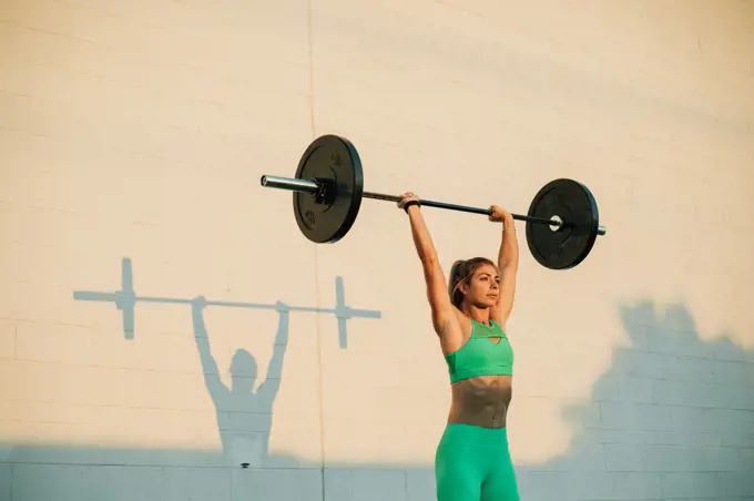 Young active woman doing overhead press with barbell in morning 