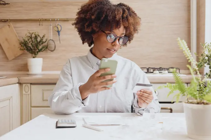 Curly woman in eye glasses pays bills in the kitchen.
