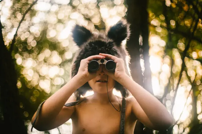 Young boy in wolf hat looks through binoculars in forest