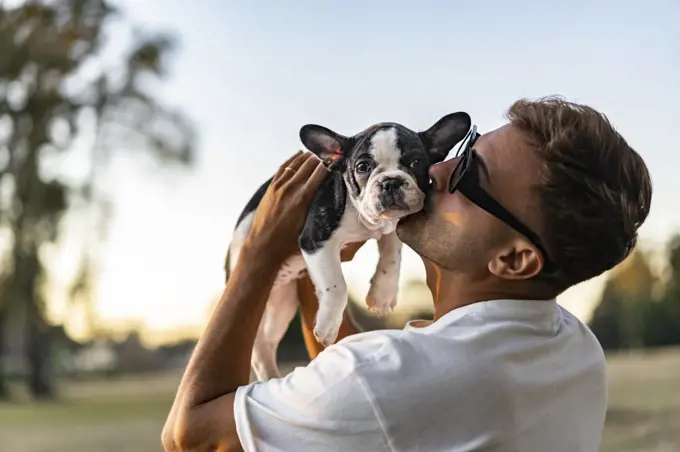 Handsome young man holding up and kissing a french bulldog.