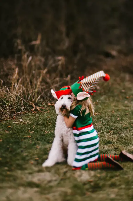Girl in elf hat kisses golden doodle dog in santa hat in grass