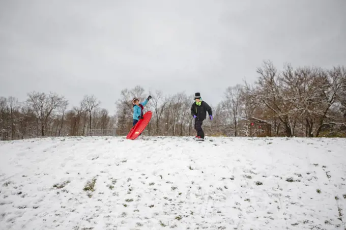 Two children stand at top of snowy hill with a sled and snowboard