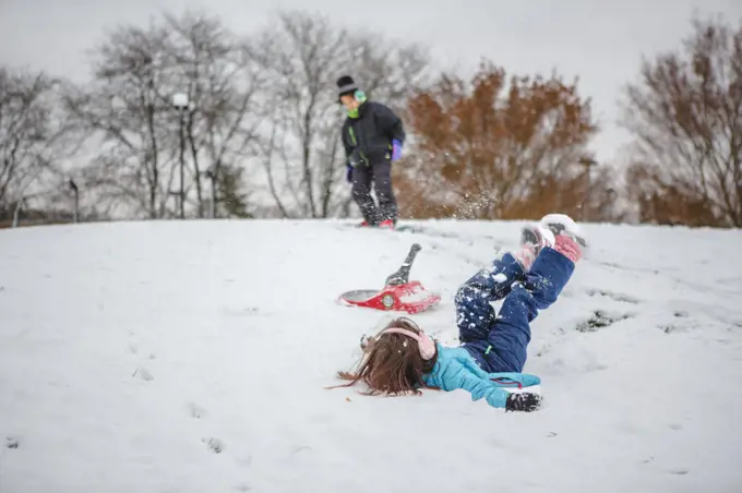 A little girl falls off sled while boy snowboards behind her