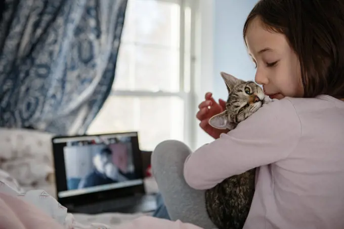 A little girl sits on bed in front of computer snuggling a kitten