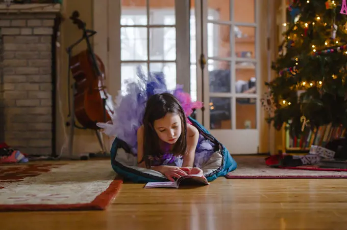 A small girl in tutu reads on floor by Christmas tree