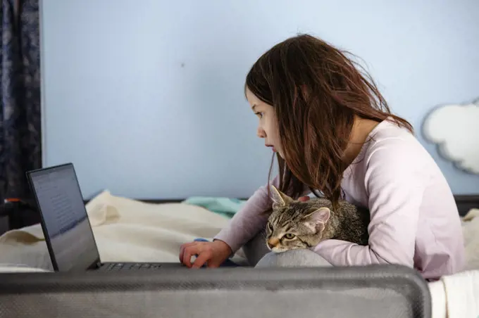 A little girl sits on bed with cat in lap looking at a computer