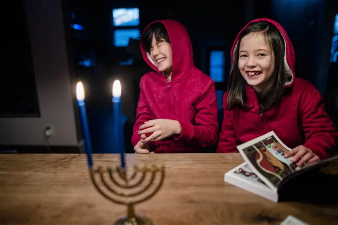 two laughing children sit at table with lit menorah at Hannukah