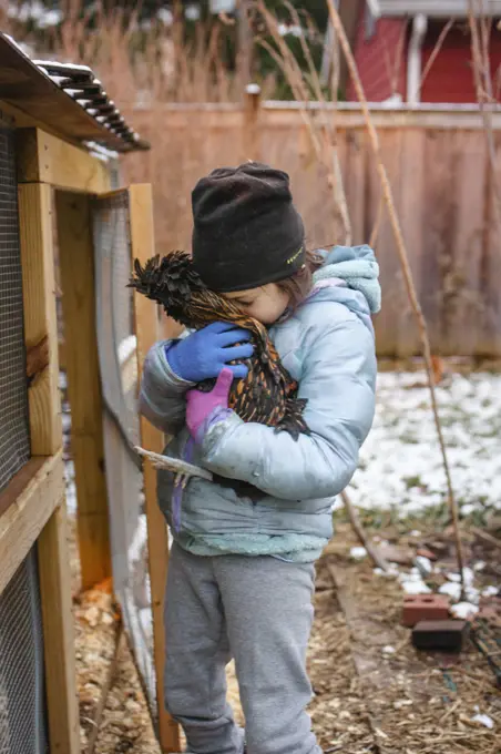 A small child nuzzles a pet chicken close to her face by chicken coop