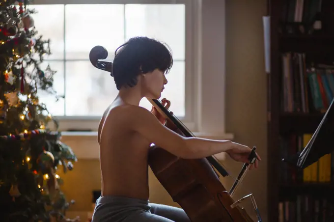 Beautiful boy practices cello by Christmas tree
