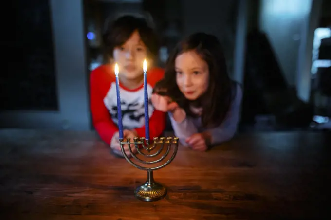 Two siblings sit at table together staring at lit candles on menorah