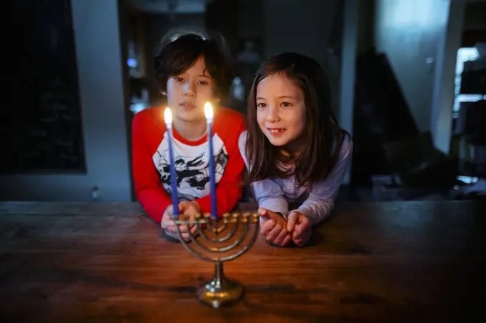 Two children sit at table together looking at lit menorah at Hannukah