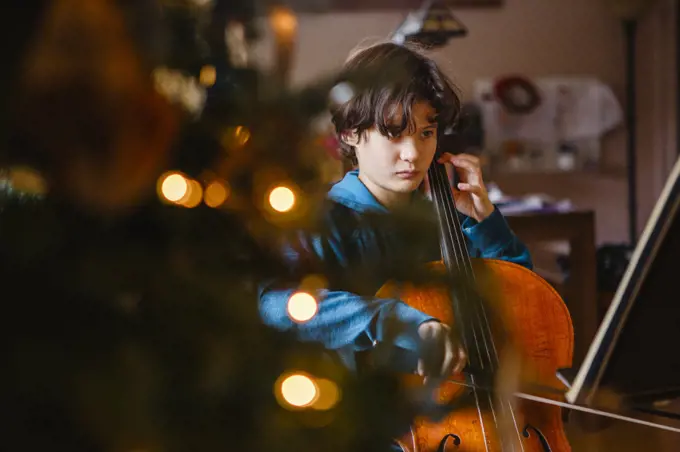A serious child plays cello by light of Christmas tree
