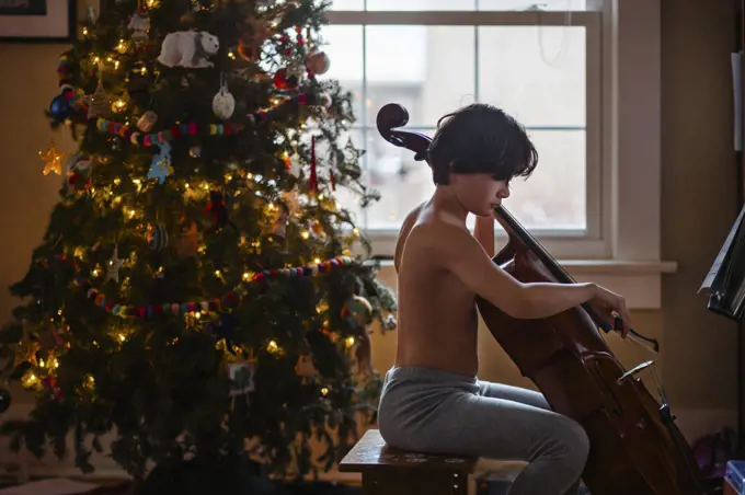 A shirtless boy plays cello by a lit Christmas tree at home