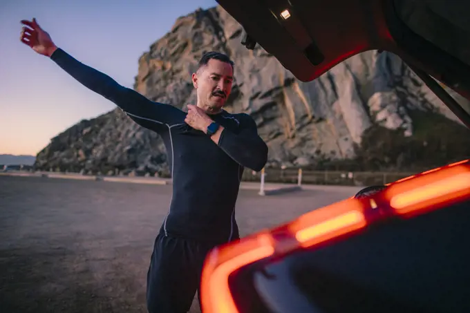 A middle aged man puts on a wetsuit next to an electric car.