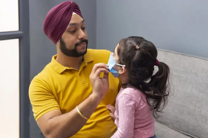 A SIKH MAN HELPING HIS LITTLE DAUGHTER TO WEAR MASK BEFORE GOING OUT
