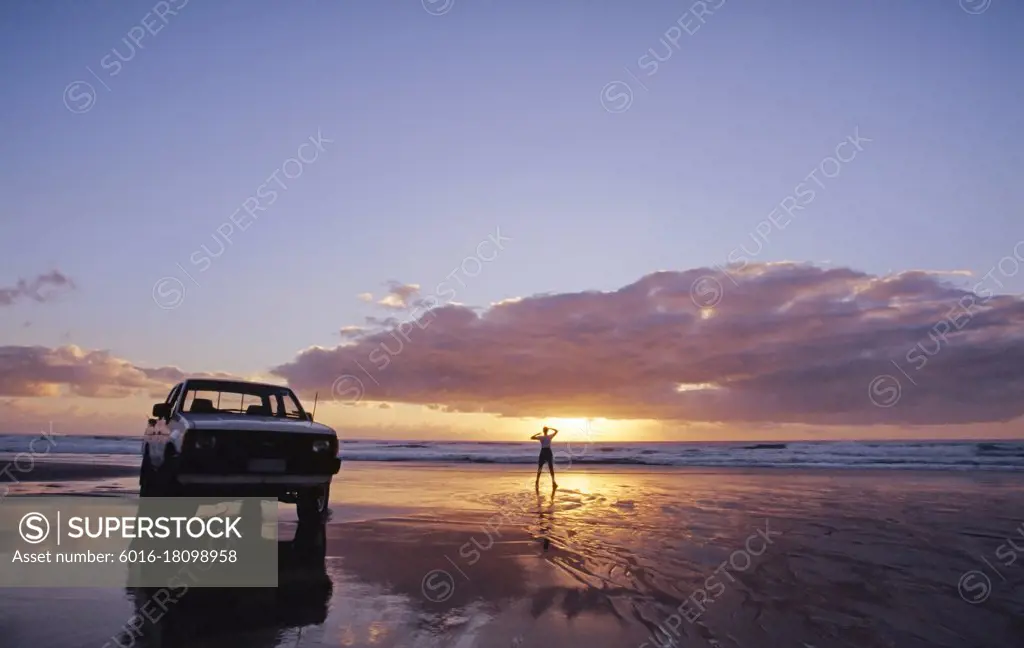 White Ute parked on the beach and lone person standing by the water at sunset