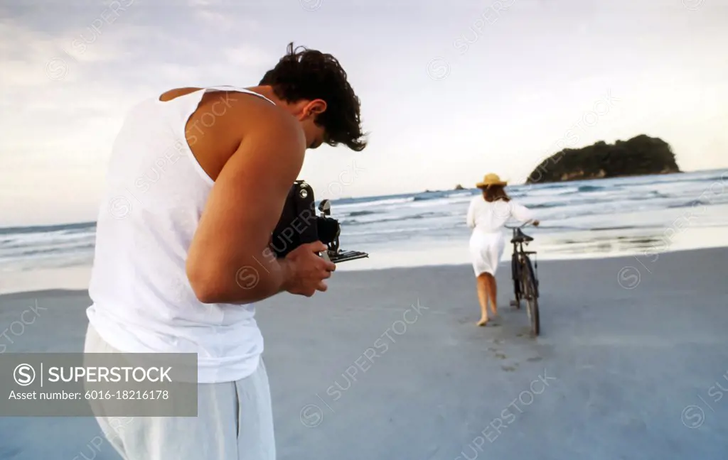 Man bent over camera taking photos of woman pushing bike in the sand at the beach